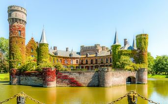 Schloss Wijnendale in Torhout mit efeubedeckten Türmen und Mauern, die sich im Wassergraben spiegeln. Rote Blumen schmücken die Steinbrücke.