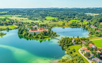 Luftaufnahme des Klosters Seeon auf einer Halbinsel in einem türkisfarbenen See, umgeben von grünen Feldern, Wäldern und einem Dorf in Bayern.