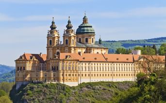 Melk Abbey perched on a rocky hill in Austria. The baroque monastery features yellow-cream facades, twin towers, and a green dome against blue sky.