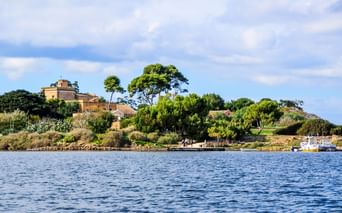Blick auf Mozia auf der Insel San Pantaleo mit historischen Gebäuden, üppigen grünen Bäumen und blauem Wasser im Vordergrund unter bewölktem Himmel.