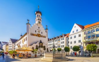 Historisches weißes Kemptener Rathaus mit Glockenturm auf Kopfsteinpflasterplatz. Brunnen im Vordergrund, bunte Gebäude und Café unter blauem Himmel.