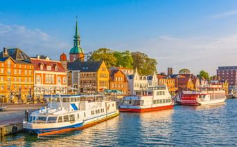 Kappeln Hafen mit weißen und roten Booten vor bunten historischen Gebäuden. Eine Kirche mit grünem Turm erhebt sich hinter gelben und orangen Fassaden.