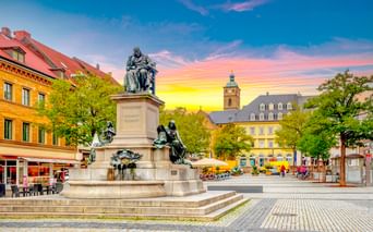 Historic market square in Schweinfurt with monument, colorful buildings, church tower, and cobblestone pavement under a vibrant sunset sky.