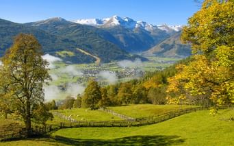 Alpental bei St. Michael im Lungau mit grünen Wiesen, Holzzäunen, Herbstbäumen und schneebedeckten Bergen unter Morgennebel.
