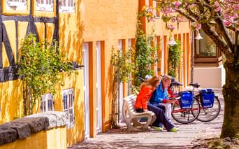 Zwei Radfahrer ruhen auf einer Bank in der Flensburger Altstadt mit Tourenrädern. Gelbe Fachwerkhäuser und ein blühender Baum bilden eine malerische Kulisse.