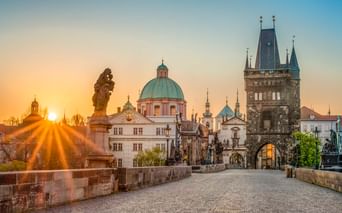 Karlsbrücke in Prag bei Sonnenaufgang mit barocken Statuen, Kopfsteinpflaster und historischen Türmen. Die Sonne erzeugt goldene Strahlen über der Altstadt.
