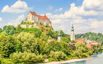 Burg Burghausen mit roten Dächern auf einem Hügel über einer Stadt mit Kirchtürmen und grünem Flussufer. Blauer Himmel mit weißen Wolken.