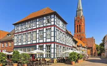 Historic market square in Nienburg with white half-timbered building and outdoor café. Red brick church with tall spire stands behind.