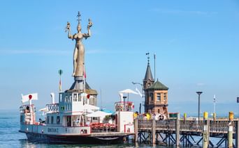 Imperia-Statue am Konstanzer Hafen mit Fahrgastschiff und historischem Turm. Die hohe rotierende Statue steht auf einem Sockel am Wasser.