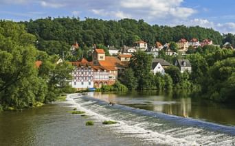 View of Melsungen on the Fulda River with a weir in the foreground. Half-timbered houses and red roofs nestle among green trees under a blue sky.