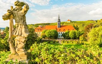 Stone statue in foreground overlooking gardens and historic buildings with orange roofs in Diesbar-Seußlitz, with vineyard-covered hills behind.