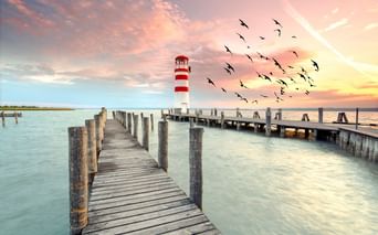 Rot-weiß gestreifter Leuchtturm auf einem Holzsteg in Podersdorf. Vögel fliegen über rosa Abendhimmel über ruhigem Wasser und Holzstegen.