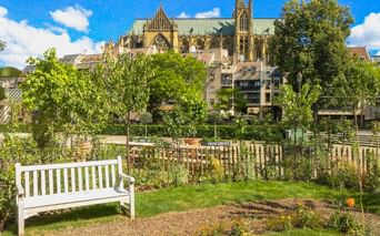 Weiße Bank im Garten mit Metzer Kathedrale hinter Wohngebäuden. Grüne Bäume und Rasen im Vordergrund unter blauem Himmel.