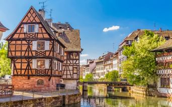 Traditionelle Fachwerkhäuser entlang eines Kanals in La Petite France, Straßburg, mit Brücke und grünen Bäumen unter blauem Himmel.
