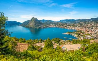Luftaufnahme von Lugano und dem Luganersee mit dem Berg Monte San Salvatore. Blauer See umgeben von Bergen und Wohngebieten unter klarem Himmel.