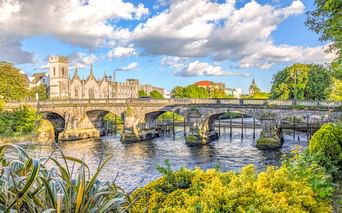 Historische steinerne Salmon Weir Bridge mit fünf Bögen über dem River Corrib in Galway, Irland. Gotische Kathedrale und üppiges Grün rahmen die Szene.