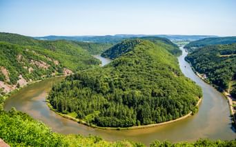Luftaufnahme der Saarschleife, einer hufeisenförmigen Flussbiegung, umgeben von dicht bewaldeten Hügeln unter blauem Himmel.