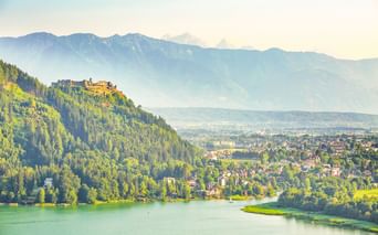Burgruine Landskron auf einem bewaldeten Hügel mit Blick auf Villach und einen türkisfarbenen See, mit Bergketten im Hintergrund bei weichem Licht.