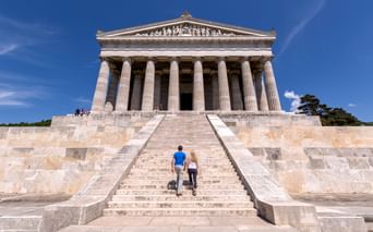 Zwei Personen steigen die Steinstufen zur Walhalla hinauf, einem neoklassizistischen Tempel mit dorischen Säulen in Bayern, unter blauem Himmel.