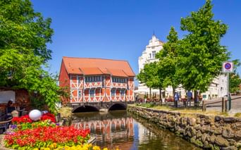 Rotes Fachwerkhaus mit orangefarbenem Dach über einem Kanal in Wismar. Bunte Blumen im Vordergrund, weißer Kirchturm dahinter, blauer Himmel.
