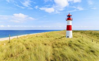 Rot-weiß gestreifter Leuchtturm auf grasbewachsenen Dünen mit Blick auf den Nordseestrand unter blauem Himmel mit weißen Wolken.