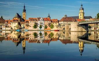 Kitzinger Altstadt mit bunten Gebäuden und Kirchtürmen, die sich im ruhigen Main spiegeln. Rechts überspannt eine Steinbrücke das Wasser.