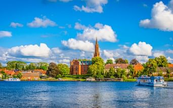 Blick über den Malchower See mit Fahrgastschiff auf blauem Wasser. Backsteinkirche mit hohem Turm ragt über Ufergebäude unter blauem Himmel.