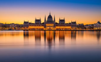 Beleuchtetes ungarisches Parlamentsgebäude am Donauufer bei Sonnenuntergang mit goldenen Lichtern, die sich im ruhigen Wasser spiegeln.
