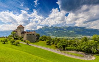 Schloss Vaduz auf einem grünen Hügel mit kurvenreicher Straße, umgeben von Bäumen. Alpenberge und dramatische Wolken im Hintergrund.