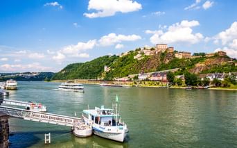 Festung Ehrenbreitstein auf einem Hügel über dem Rhein in Koblenz. Fähren und Passagierschiffe auf dem Wasser, blauer Himmel mit Wolken.