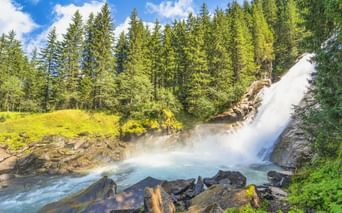 Krimmler Wasserfälle stürzen über Felsen in türkisfarbenes Becken, umgeben von grünem Wald und Wiesen unter blauem Himmel mit aufsteigendem Nebel.