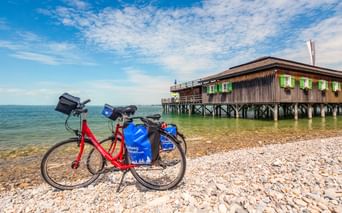 Rotes Tourenrad mit blauen Taschen am Kieselstrand in Bregenz. Hölzerne Badehütte auf Stelzen mit grünen Fensterläden ragt in den Bodensee.