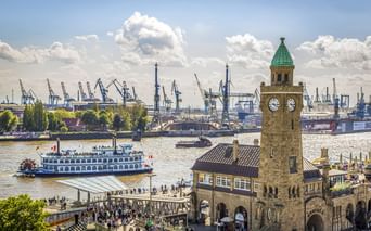 St. Pauli Landungsbrücken in Hamburg mit Uhrturm, Raddampfer auf der Elbe und Hafenkränen im Hintergrund unter bewölktem Himmel.