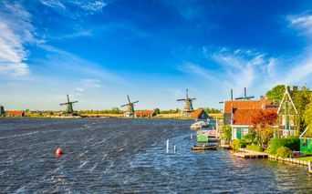 Drei historische Windmühlen entlang eines Wasserwegs in Zaanse Schans mit traditionellen niederländischen Häusern mit orangefarbenen Dächern unter blauem Himmel.