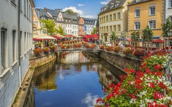 Steinbrücke mit roten und weißen Blumen über einem Kanal in Saarburgs Altstadt, mit bunten historischen Gebäuden und roten Sonnenschirmen.