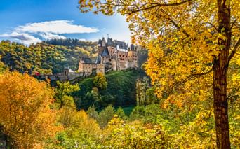 Burg Eltz thront auf einem Hügel, umgeben von dichtem Wald mit goldenem Herbstlaub unter blauem Himmel mit weißen Wolken.