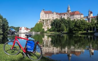 Rotes Fahrrad mit blauen Taschen auf Wiese an der Donau. Schloss Sigmaringen thront auf einem Felsen über dem Wasser und spiegelt sich in der ruhigen Oberfläche.