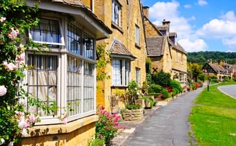Reihe honigfarbener Steinhäuser mit blühenden Gärten entlang einer Dorfstraße in den Cotswolds unter blauem Himmel mit weißen Wolken.