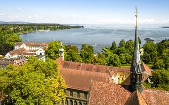 Rote Ziegeldächer und Kirchturm mit Blick auf den Bodensee mit grünen Bäumen, Segelbooten auf dem Wasser und fernem Ufer unter blauem Himmel.