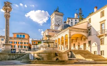Historischer Platz in Udine mit verziertem Brunnen, weißem Uhrenturm mit blauer Kuppel, Arkadenloggia und bunten Gebäuden unter blauem Himmel.