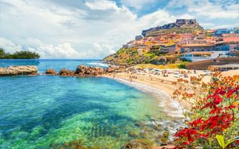 Sandstrand mit klarem türkisfarbenem Wasser in Castelsardo, Sardinien. Bunte Häuser erstrecken sich den Hügel hinauf, gekrönt von einer Festung.
