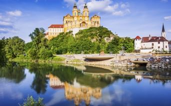Gelbes barockes Stift Melk auf grünem Hügel, gespiegelt in der ruhigen Donau, mit weißer Kirche am rechten Ufer sichtbar.