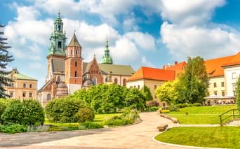 Wawel-Kathedrale in Krakau mit markanten grünen Kupfertürmen und roten Backsteinmauern, umgeben von gepflegten Rasenflächen und Gärten unter blauem Himmel.
