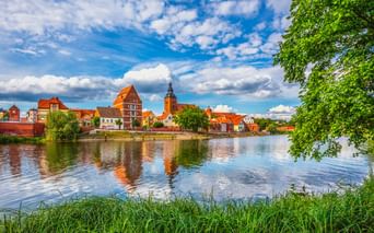 Blick auf Havelberg über den Fluss mit historischen Gebäuden mit roten Dächern und Kirchturm, die sich im ruhigen Wasser spiegeln.