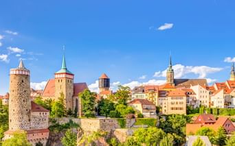 Panoramablick auf Bautzens Altstadt mit mittelalterlichen Türmen, Kirchtürmen und bunten Gebäuden auf einem Felsenhügel unter blauem Himmel.