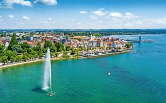Luftaufnahme von Friedrichshafen am Bodensee mit hohem Springbrunnen im türkisfarbenen Wasser, historischer Altstadt, Kirchturm und Fahrgastschiff.