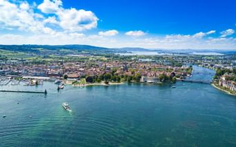 Luftaufnahme von Konstanz und Kreuzlingen am Bodensee mit historischen Gebäuden, einem Hafen und Booten auf türkisfarbenem Wasser unter blauem Himmel.