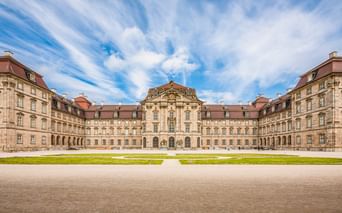 Symmetrisches Barockschloss mit roten Ziegeldächern und Sandsteinfassade um einen Innenhof mit grünem Rasen unter dramatischem Wolkenhimmel.