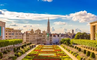 Formale Gärten am Mont des Arts in Brüssel mit bunten geometrischen Blumenbeeten, gestutzten Hecken und Stadtsilhouette mit Kirchturm unter blauem Himmel.