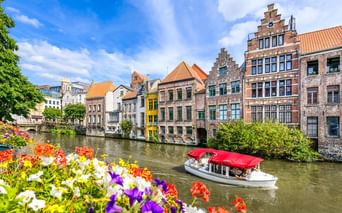 Touristenboot mit rotem Dach auf Kanal in Gent, gesäumt von bunten historischen Gebäuden. Bunte Blumen rahmen den Vordergrund.